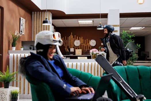 Woman enjoys online connectivity in hotel lobby, using digital laptop for communication and relaxation. Female traveler dressed in skiing gear sits with minicomputer in lounge area for winter holiday.