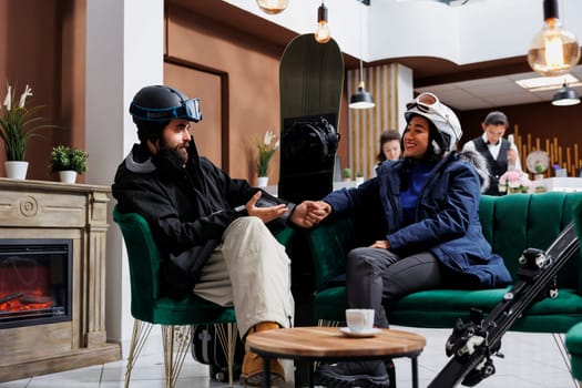 Couple dressed in warm winter gear sitting and talking on cozy sofa in hotel lobby. Excited man and woman wearing ski goggles and winter jackets are holding hands, ski equipment leaning against couch.