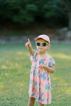 Little girl in sunglasses stands in a clearing pointing her finger into the distance. High quality photo