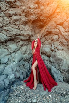 Red dress rocks woman. A blonde with flowing hair in a long flowing red dress stands near a rock of volcanic origin. Travel concept, photo session at sea.