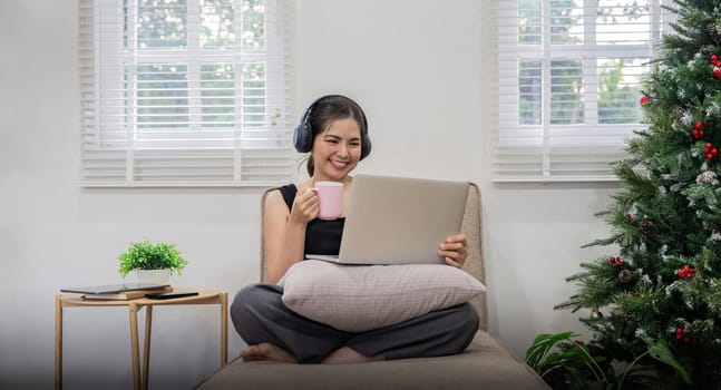 Pretty young woman asian listening to music with laptop relaxing and drinking tea or coffee while sitting on sofa at home.