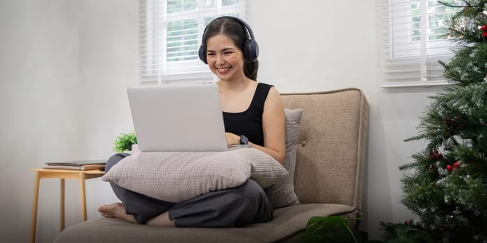 Young Asian woman listening music from headphones and sitting on sofa in living room.