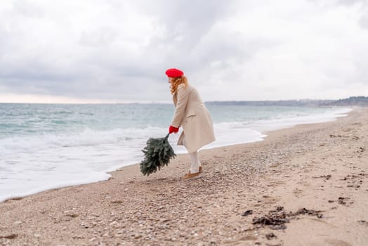 Redhead woman Christmas tree sea. Christmas portrait of a happy redhead woman walking along the beach and holding a Christmas tree in her hands. She is dressed in a light coat and a red beret
