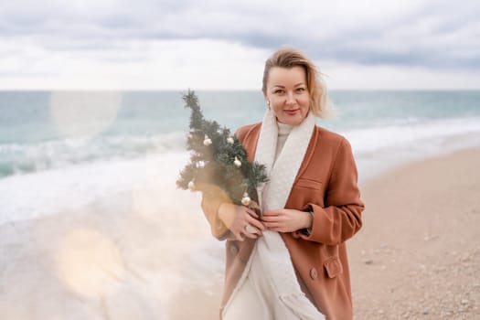 Blond woman Christmas sea. Christmas portrait of a happy woman walking along the beach and holding a Christmas tree in her hands. She is wearing a brown coat and a white suit