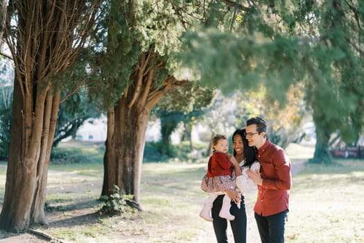 Smiling dad hugging mom from behind with a little girl in her arms standing on a path near a tree. High quality photo