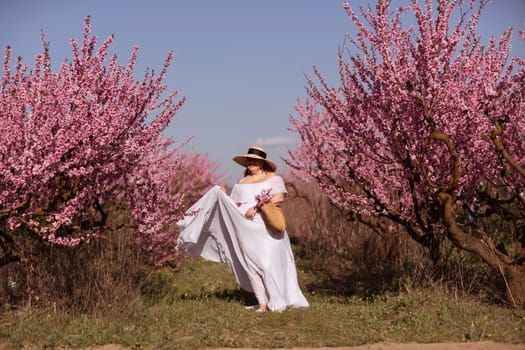 Woman blooming peach orchard. Against the backdrop of a picturesque peach orchard, a woman in a long white dress and hat enjoys a peaceful walk in the park, surrounded by the beauty of nature