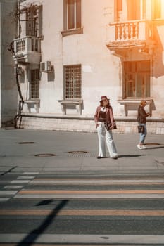 Woman city road crossing. Stylish woman in a hat crosses the road at a pedestrian crossing in the city. Dressed in white trousers and a jacket with a bag in her hands