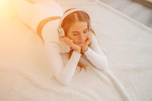 Top view portrait of relaxed woman listening to music with headphones lying on carpet at home. She is dressed in a White tracksuit