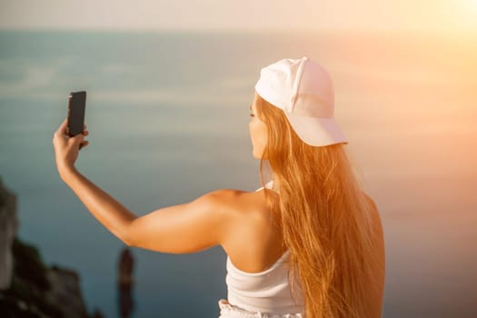 Selfie woman sea. The picture depicts a woman in a cap and tank top, taking a selfie shot with her mobile phone, showcasing her happy and carefree vacation mood against the beautiful sea backgroun.