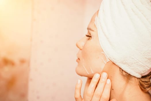 Young beautiful woman using a moisturizing facial mask after taking a bath. Pretty attractive girl in a towel on her head stands in front of a mirror in a home bathroom. Daily hygiene and skin care.