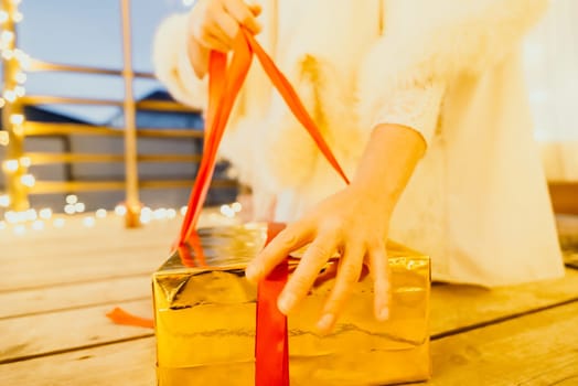 A woman in a white dress is holding a gold box with a red ribbon. She is wearing a crown on her head. The scene takes place in a room with a door and a window. The woman appears to be opening the gift box.