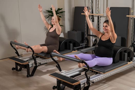 Two pregnant women do Pilates exercises on a reformer. Yoga class for pregnant women