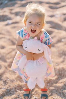 Little girl screams while standing on the beach hugging a toy hare. Top view. High quality photo