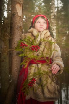 Portrait of Teen girl in coat, red sash and branch of fir tree with bright berries in cold winter day in forest. Medieval peasant girl with firewood. Photoshoot in stile of Christmas fairy tale