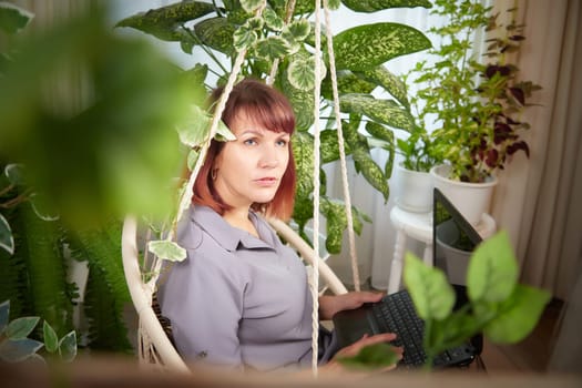 A girl with a laptop and notebook is working on a swing chair in the winter garden. A middle-aged business woman. Psychologist in the office with flowers