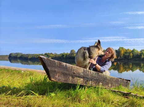 Adult girl with shepherd dog taking selfie on water of river, lake with old boat. Middle aged woman and big pet on nature. Friendship, love, fun, hugs