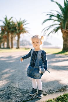 Little smiling girl stands on the road near palm trees with a toy in her hand. High quality photo