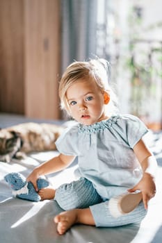 Little girl with a brush in her hand sits next to a soft toy on the bed. High quality photo