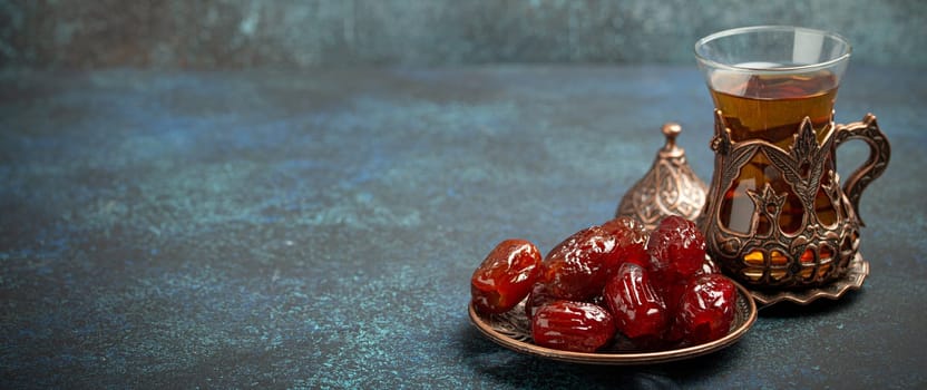 Breaking fasting with dried dates during Ramadan Kareem, Iftar meal with dates and Arab tea in traditional glass, angle view on rustic blue background. Muslim feast, space for text.