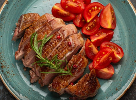 Delicious roasted sliced duck breast fillet with golden crispy skin, with pepper and rosemary, top view on ceramic blue plate served with cherry tomatoes salad, rustic concrete rustic background.