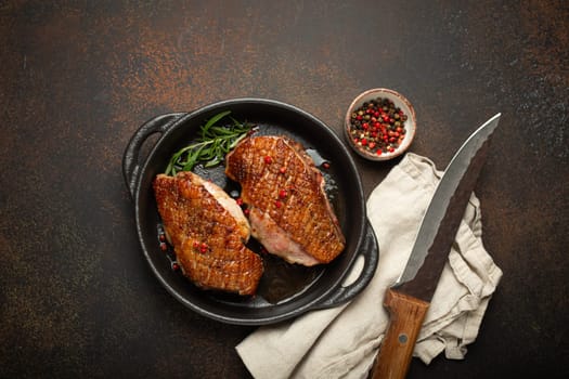 Two roasted duck breast fillets with crispy skin, with pepper and rosemary, top view in black cast iron pan with knife, dark brown concrete rustic background.