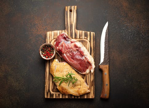 Two raw uncooked duck breast fillets with skin, seasoned with salt, pepper, rosemary top view on wooden cutting board with knife, dark brown concrete rustic background.