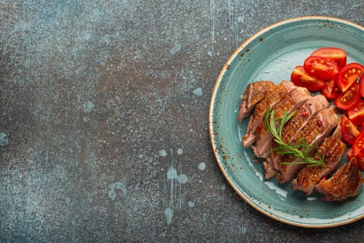 Delicious roasted sliced duck breast fillet with golden crispy skin, with pepper and rosemary, top view ceramic blue plate served with cherry tomatoes salad, concrete rustic background, copy space.
