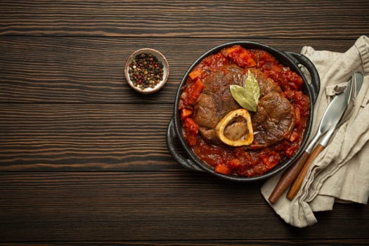Traditional Italian dish Ossobuco all Milanese made with cut veal shank meat with vegetable tomato sauce served in black casserole pan top view on rustic brown wooden background, copy space.