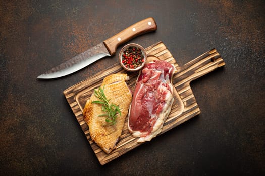 Two raw uncooked duck breast fillets with skin, seasoned with salt, pepper, rosemary top view on wooden cutting board with knife, dark brown concrete rustic background.