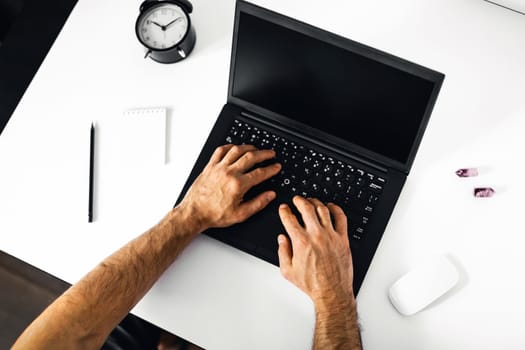 The man working at home on the remote. A black-and-white workplace with a laptop, alarm clock, notebook and men's hands.