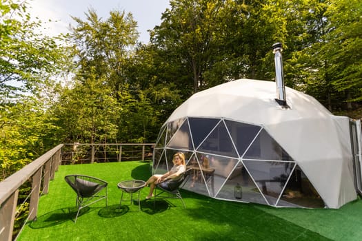 Young woman resting on sunbed on the wooden terrace near the modern house with panoramic windows near pine forest. Concept of solitude and recreation on nature.