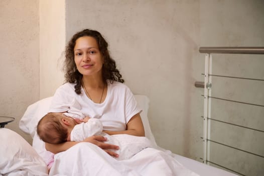 Beautiful Latin American woman, young loving caring mother in white pajamas, breastfeeding her baby in cozy home bedchamber, smiling and confidently looking at the camera. Maternity leave lifestyle