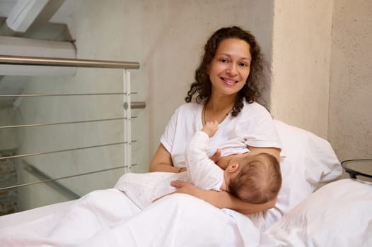 Latin American young beautiful woman mother breastfeeds her baby, holding him in her arms while waking up in the morning, sitting on the bed and smiling from happiness, looking confidently at camera