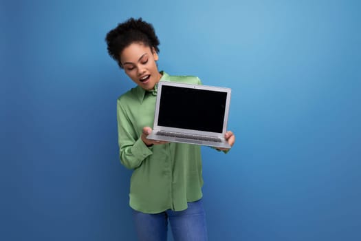 young pretty female model with curly black hair dressed in a green blouse and jeans holds a laptop screen forward with a mock up for advertising.