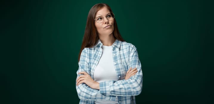 Young European woman dressed in a plaid shirt and jeans thinking on a dark green background.