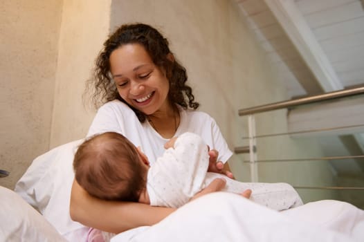 Beautiful multi ethnic woman, happy loving young mother smiles while connects with her newborn baby, holds him in her arms and breastfeeds him, sitting on the bed in white home bedchamber interior