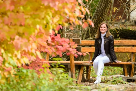 A young beautiful girl sits on a bench in an autumn park and looks happily into the frame a