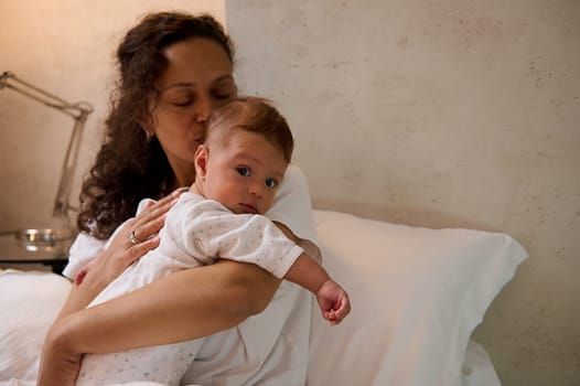 Portrait of authentic beautiful Latin American woman in white pajamas, holding and gently cuddling her little baby boy, sitting together on the bed in cozy bedroom interior. Maternity leave lifestyle