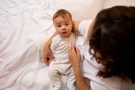 Portrait of authentic little baby boy in the hands of his mother, sitting together on the bed in cozy bedroom interior. Maternity leave lifestyle, infancy, babyhood. Motherhood. Single parenting