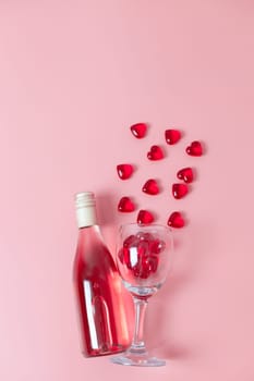 One bottle of wine and a glass with glass hearts pouring out of it lies in the center on a pink background, flat close-up. The concept of love and the holiday of Valentine's Day.