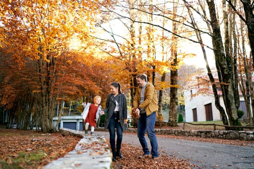 Dad throws fallen leaves at mom with little girl holding hands in the park. High quality photo