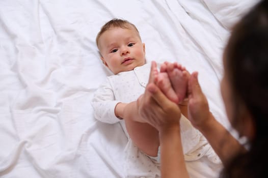 Beautiful newborn baby boy, looks at the camera while his loving caring young adult mother strokes and kisses his little feet and tiny toes. Family relationships. Connection. Infancy and motherhood