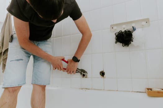 One young Caucasian recognizable guy with a brunette unscrews the pipe from the faucet with an adjustable wrench in a white tiled wall in the bathroom at home, close-up side view.Step by step.