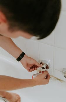 One young Caucasian brunette guy wraps white tow on a faucet nut sticking out of the wall, standing bent over in the bathroom, close-up view from above. Step by step.