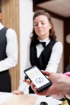 Female receptionist at front desk providing assistance to tourist using mobile phone for nfc payment. Image showcasing hotel concierge holding pos terminal for traveler to pay with smartphone.