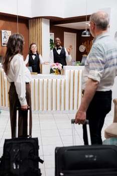 Caucasian elderly couple enters hotel and approaches front desk for reservation assistance from multiethnic employees. Professional receptionists greeting retired seniors with smiles.