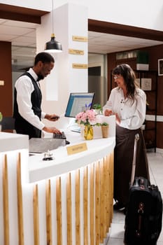 African american receptionist assists old woman with hotel check-in verifying identity and retrieving reservation details. Male concierge holds and checks passport of female tourist. Side-view shot.