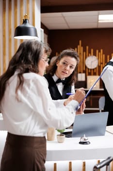 Professional concierge assists retired senior woman at hotel lobby, ensuring smooth check-in process. Receptionist provides friendly service to elderly lady traveler as she signs reservation forms.