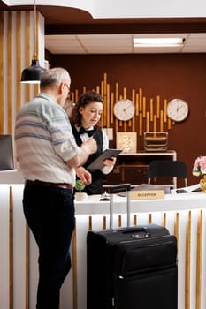 Elderly male traveller checks in at registration counter of hotel lobby, eager to begin enjoyable holiday. Retired senior man fills out reservation documents with help from kind female receptionist.