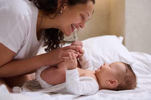 Close up portrait of happy young adult woman, a loving caring mother holding legs of her newborn baby near her face, smiling and looking tenderly at him while kissing him and spending time in bedroom